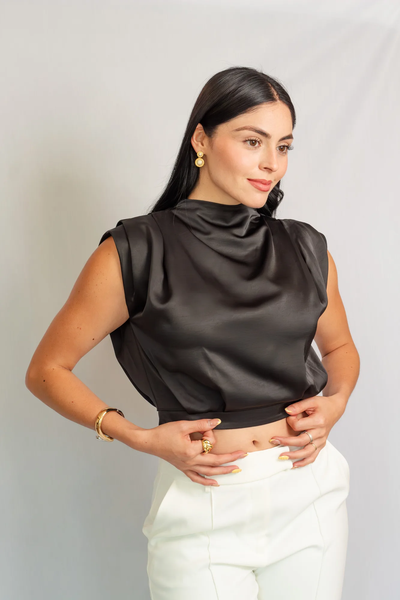 Modelo con blusa corta de satín negro de cuello alto drapeado y pantalón blanco.